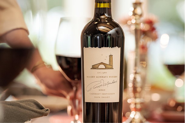 Bottle of 2021 Nap Valley Cabernet Sauvignon on a table with glasses of wine and hands behind it