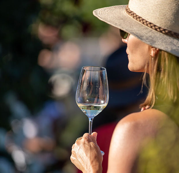 A woman in profile wearing a wide-brimmed straw hat and sunglasses, holding a wine glass with white wine in a sunlit outdoor setting.