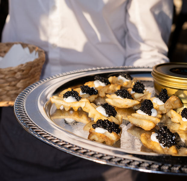 A close-up of a silver serving tray holding small appetizers topped with black caviar, held by a person in a white shirt. A woven basket and a gold-lidded container are partially visible in the background.