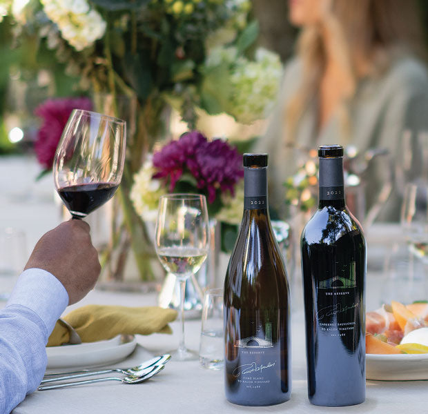A bottle each of Robert Mondavi Winery The Reserve Cabernet Sauvignon and Fume Blanc, on a dining table with wine glasses, a hand holding a glass of red wine, and blurred flowers in the background. 